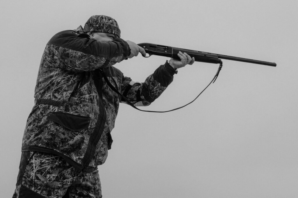 Home Black and white image of a hunter aiming a rifle, dressed in camouflage, against a cloudy sky.