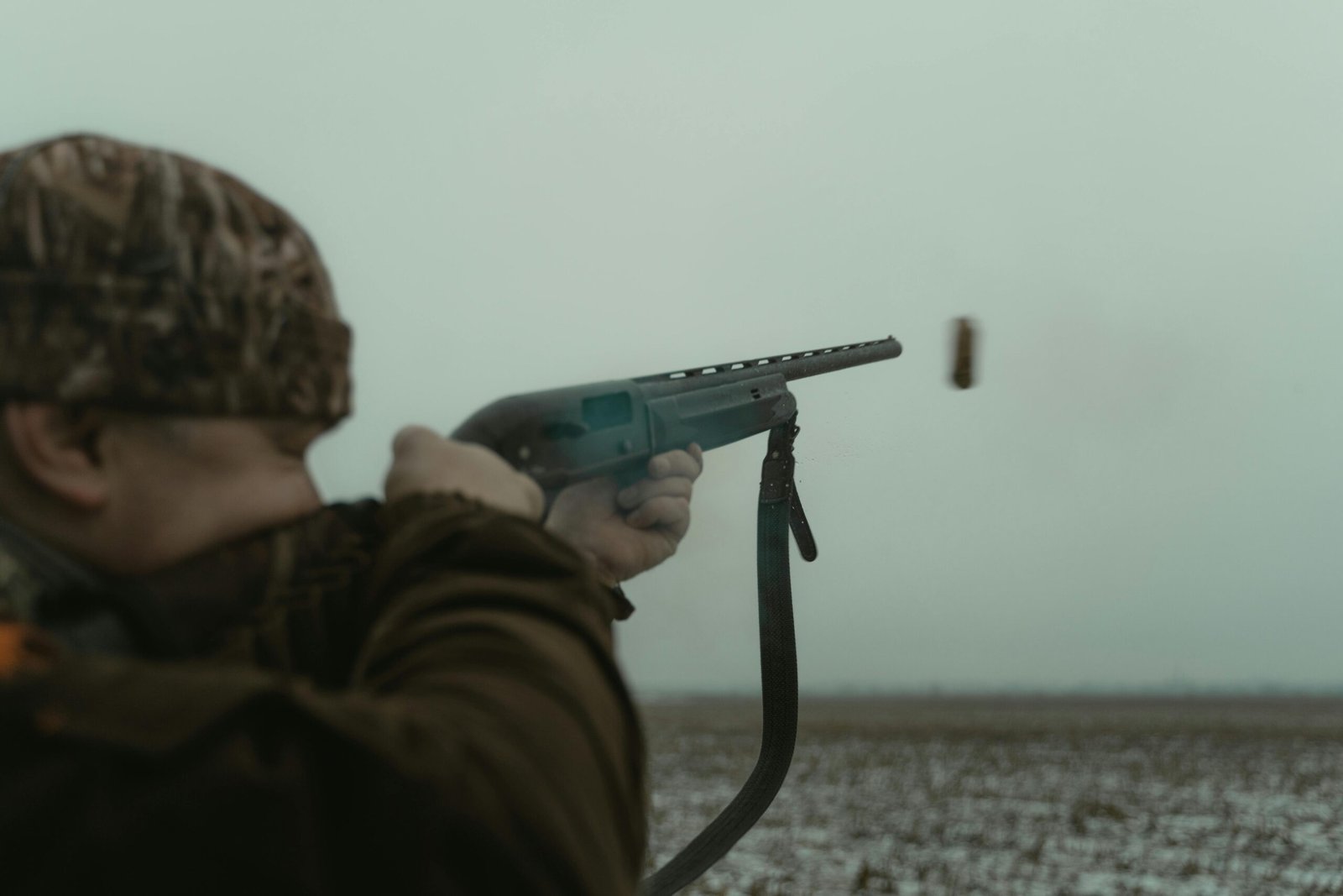 Adult male aiming a shotgun in a rural setting with camouflage clothing.